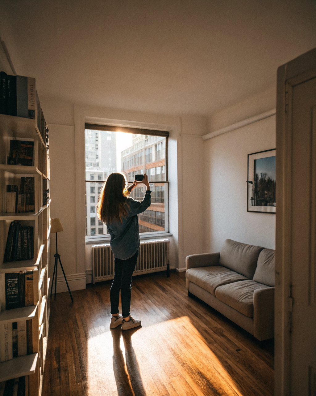 A woman holding up her smartphone to photograph her small apartment living room, with morning light coming through a window partially blocked by a bookshelf